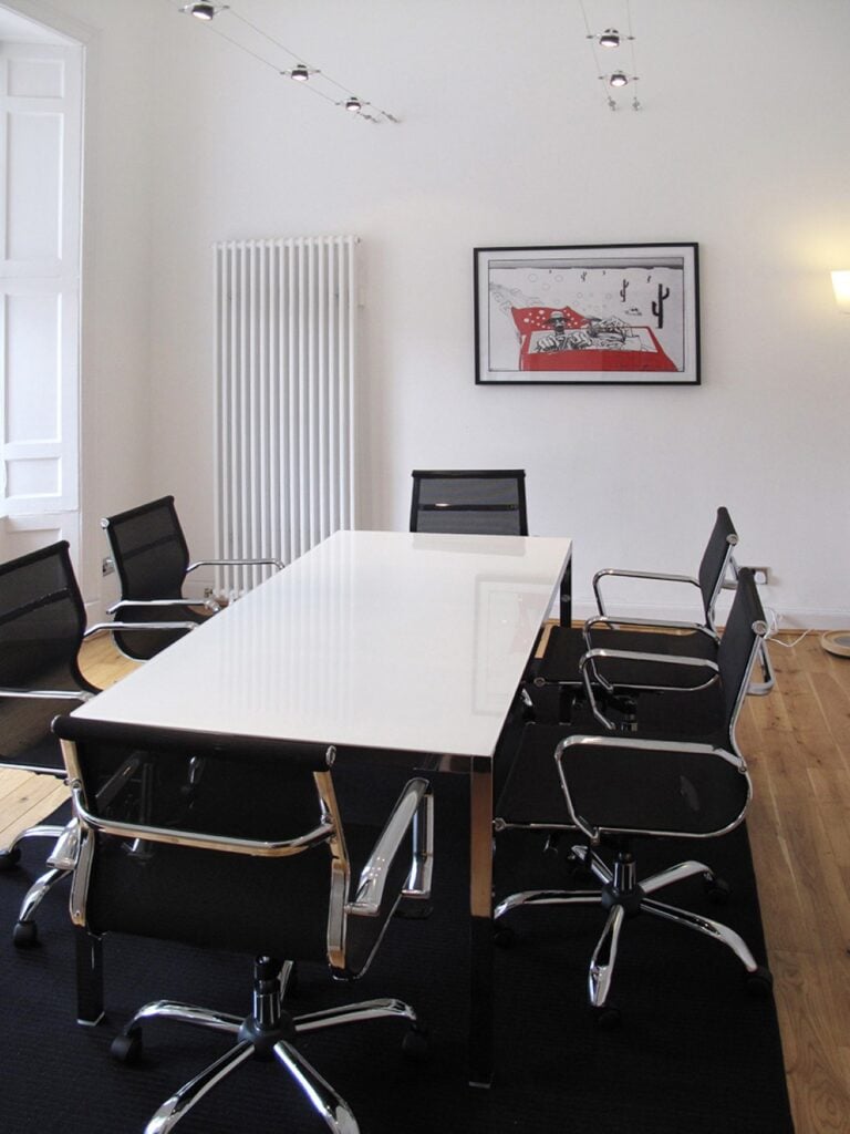 A modern conference room by 44 Unlimited features a long white table surrounded by six black office chairs. A contemporary art piece hangs on the white wall above a radiator, while wooden flooring and track lighting enhance its sleek design.