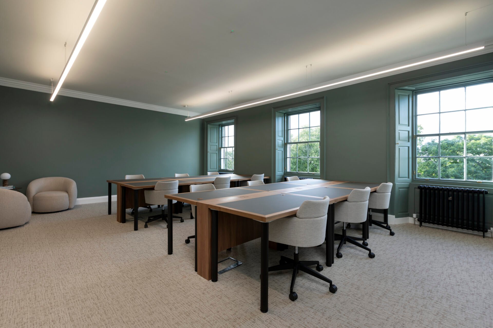 Discover a modern minimalist office space at 22 Queen Street, boasting wooden desks and beige chairs. This tranquil room features green walls, large windows with lush views, contemporary lighting, and two cozy beige armchairs nestled in the corner.