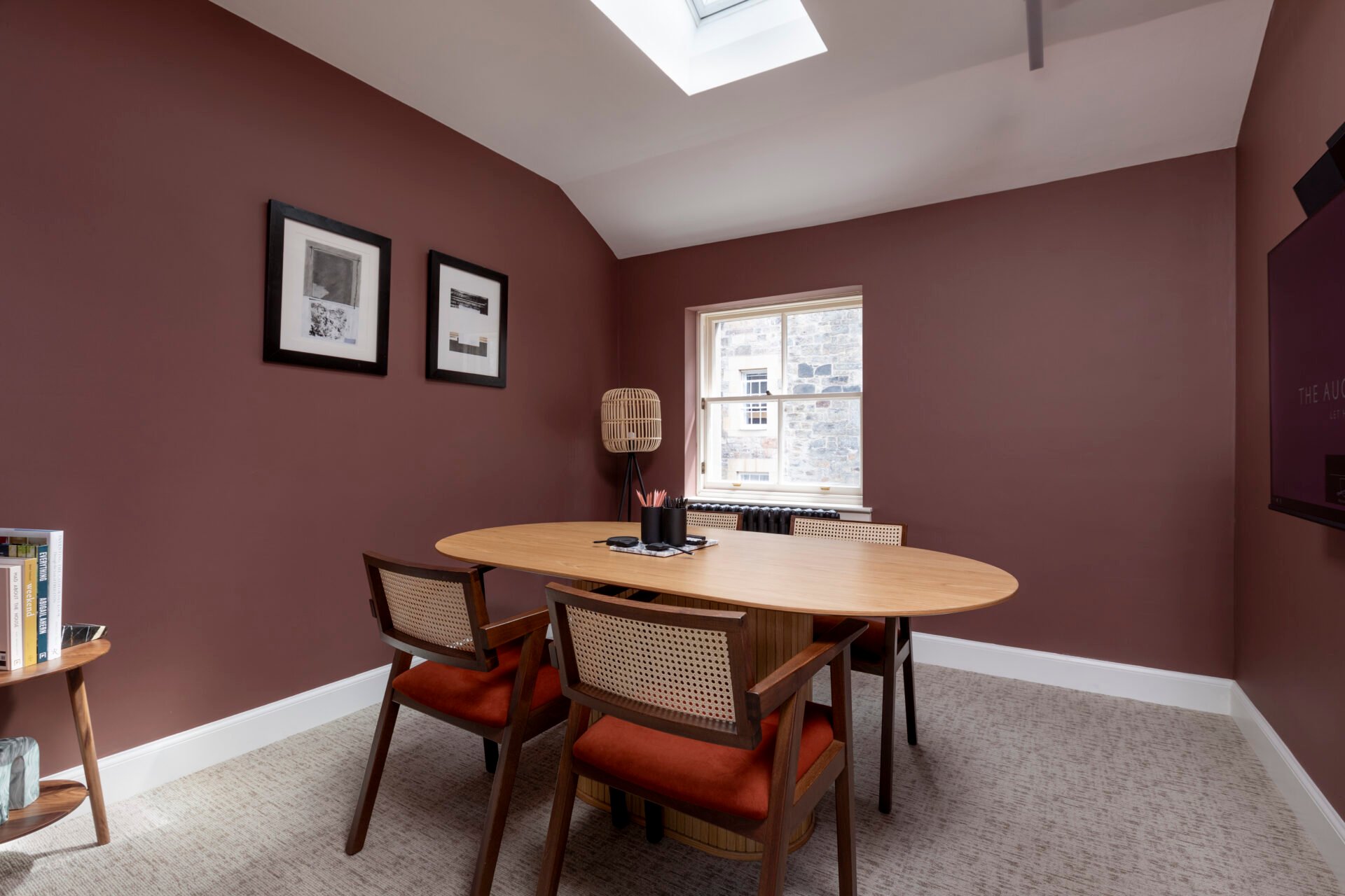 Located at 22 Queen Street, this modern office space boasts a brown oval table with four wooden chairs featuring woven backs and red cushions. The brown walls display two framed pictures, while a window and skylight fill the room with natural light. A bookshelf sits in the corner.