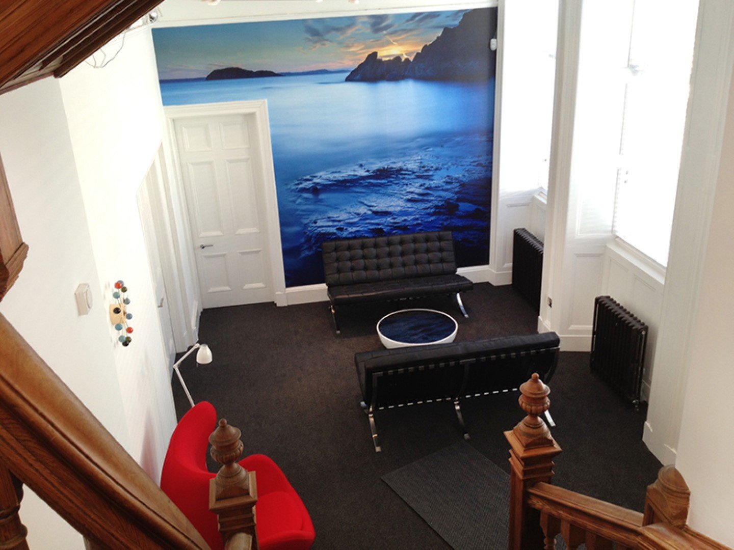 An office lounge area with two black sofas and a red chair arranged around a circular coffee table from 93 Unlimited. A large mural of a seascape covers the wall. The space is well-lit by natural light from two large windows, while a wooden staircase is partially visible.