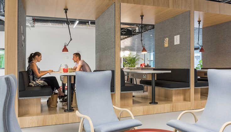 People at Lochrin Square are seated in a modern office space with wooden booths and red pendant lights. Two individuals sit across from each other, deep in conversation. Neutral-colored chairs and decor provide an inviting foreground.