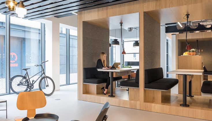 A modern office space at Lochrin Square boasts wooden cubicles with cushioned seating and tables. A person works on a laptop, while a bicycle rests by large windows. Overhead lights illuminate the contemporary chairs scattered throughout.