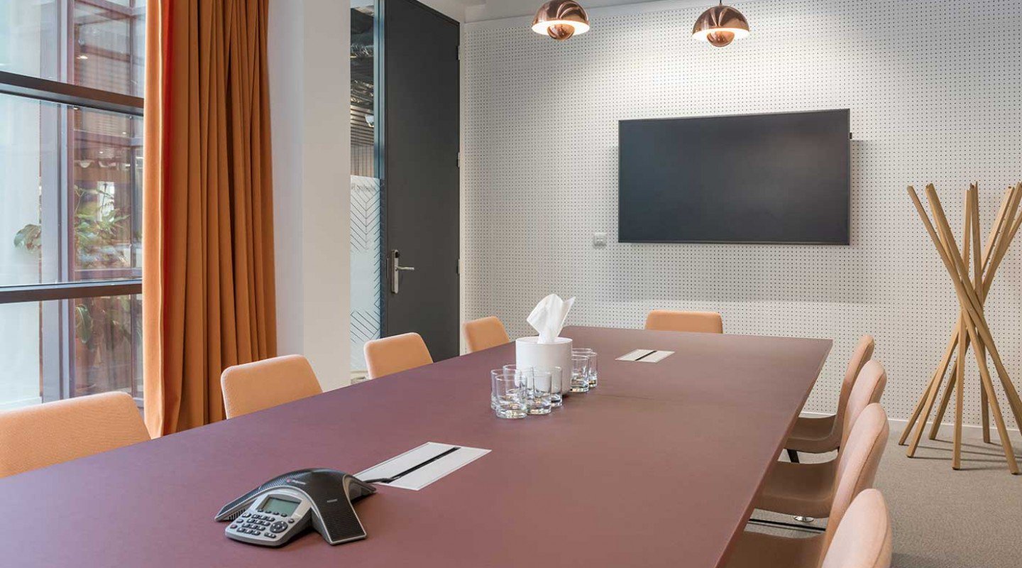 Located in Lochrin Square, this modern meeting room features a long maroon table, six beige chairs, and a wall-mounted flat-screen TV. A conference phone and glasses sit on the table while orange curtains and a coat rack add subtle touches to its minimalist, professional feel.