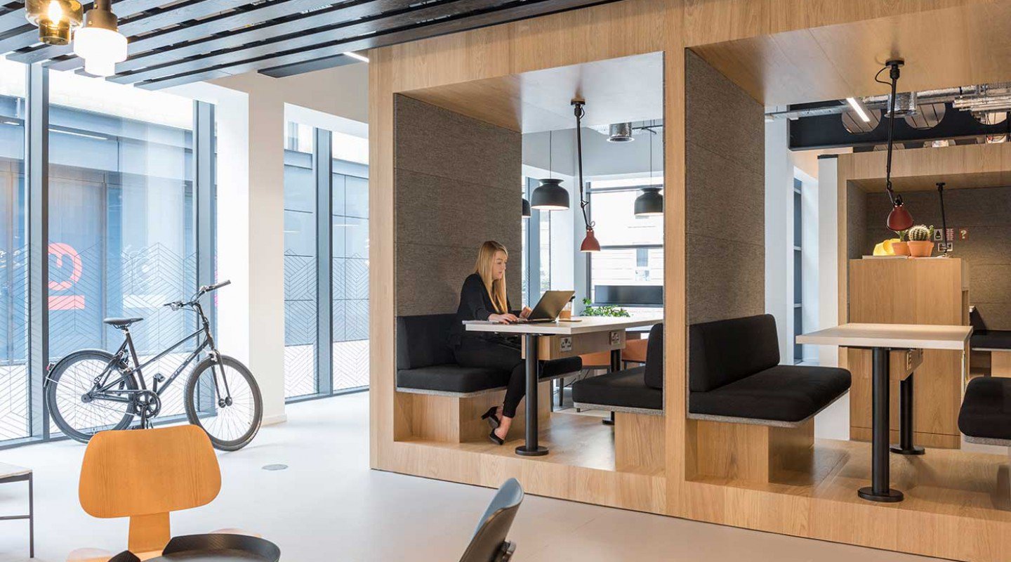 In the contemporary, minimalist office at Lochrin Square, a woman works diligently on her laptop within the wooden cubicles. Nearby, a bicycle rests against a window and pendant lights elegantly hang from the ceiling.