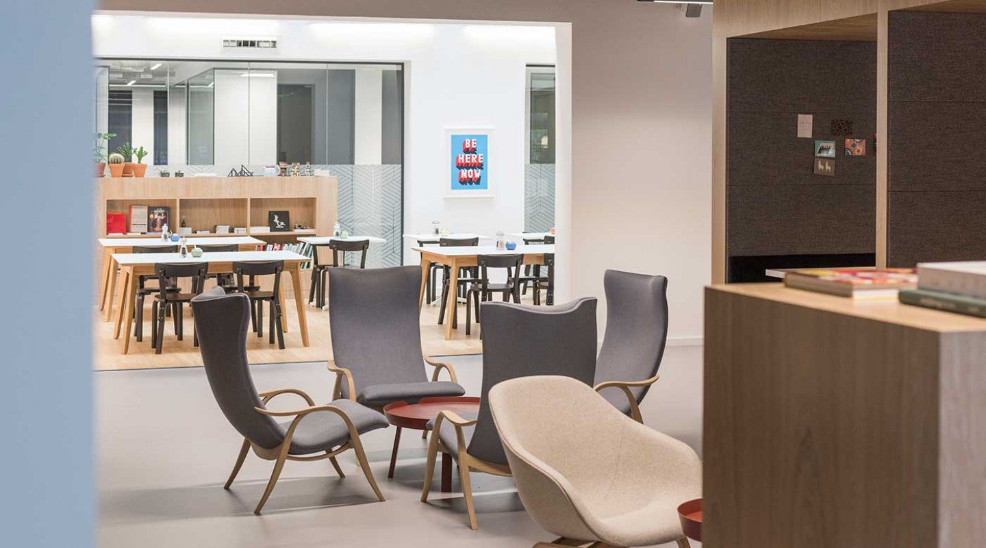 Located at Lochrin Square, this modern office space features stylish chairs in the foreground and a meeting area in the background, complete with tables and chairs. A wall poster reads We Are Here Now. The setting is clean and minimalist, with light wood and neutral tones.