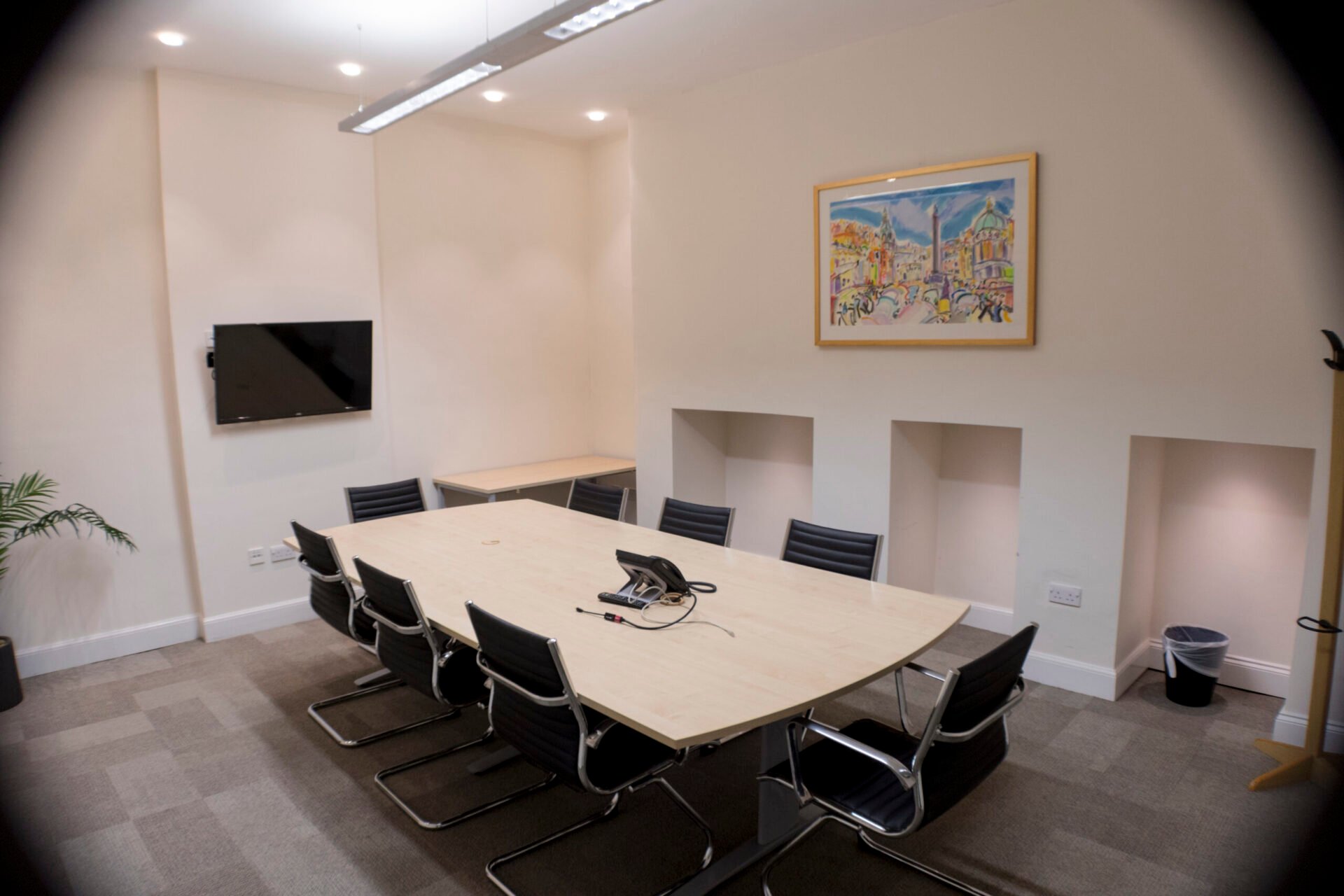 In Edinburgh, a modern conference room features a long wooden table with eight black leather chairs. A flat-screen TV adorns the wall, while a vibrant painting adds color. The space is complete with a phone on the table and a potted plant in the corner, exuding style and functionality.